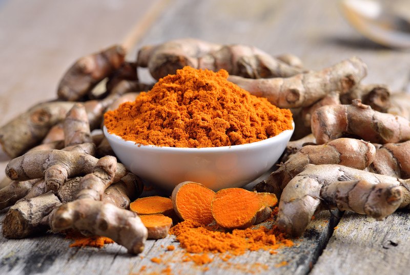 This photo shows a bowl of turmeric powder, a sliced turmeric root, and several uncut turmeric roots lying on a wooden table.