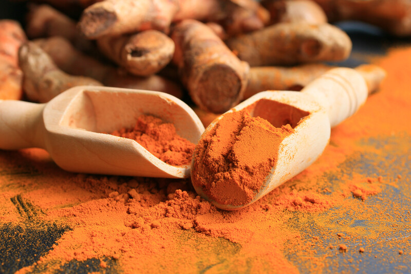 Two wooden scoop full of turmeric powder with blurred image of fresh turmeric at the back.