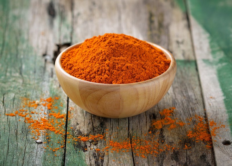 a wooden bowl full of turmeric  powder resting on aged-wooden surface, with loose turmeric powder beside it