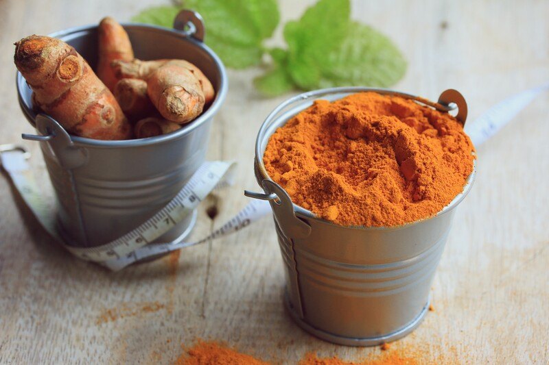 a couple of small tin pail on a wooden surface, one with turmeric powder and the other full of fresh turmeric; with white tape measure and fresh basil life at the back