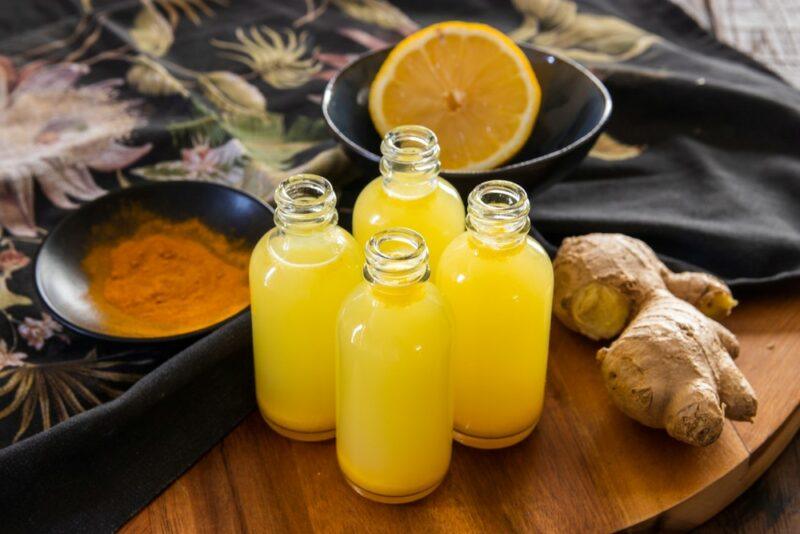 Four small bottles containing turmeric shots, next to some turmeric, orange, ginger, and honey