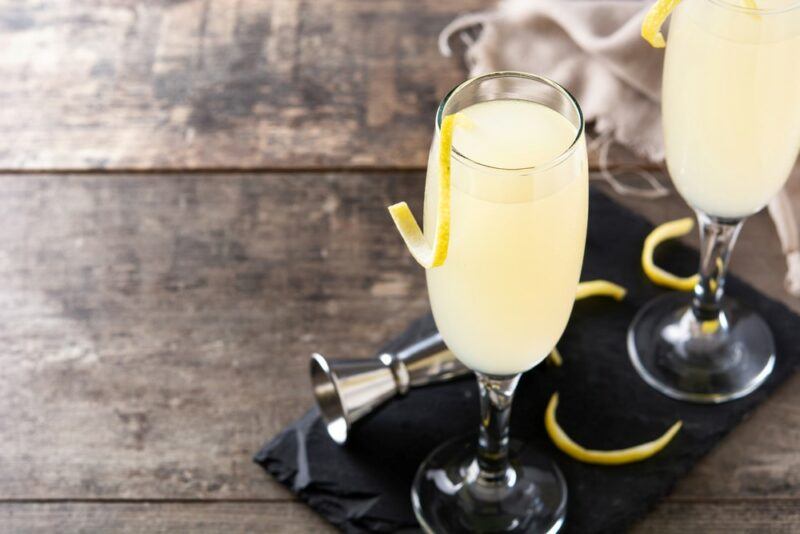 A black plate on a wooden table with two French 75 cocktails, a jigger, and lemon twists