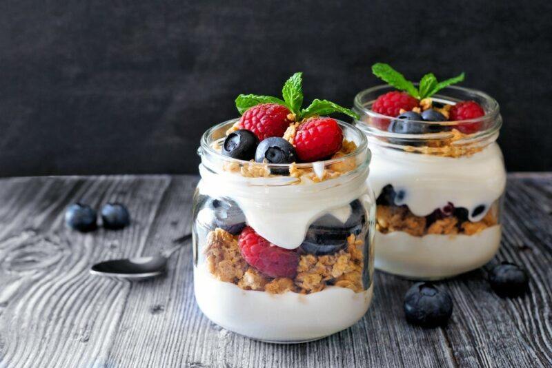 A wooden table with two jars of Greek yogurt parfait that include layers of yogurt, berries, and granola