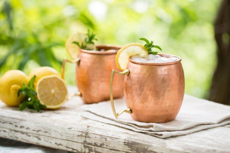 Two copper mugs containing a Moscow mule cocktails, with lemons next to them and greenery outside