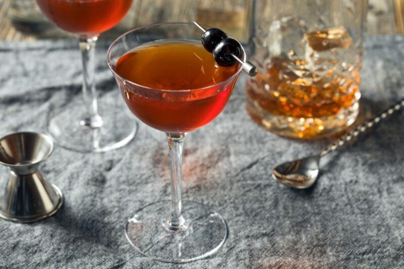 Two glasses containing Rob Roy cocktails on a bar, with a glass of whiskey in the background and some bar tools