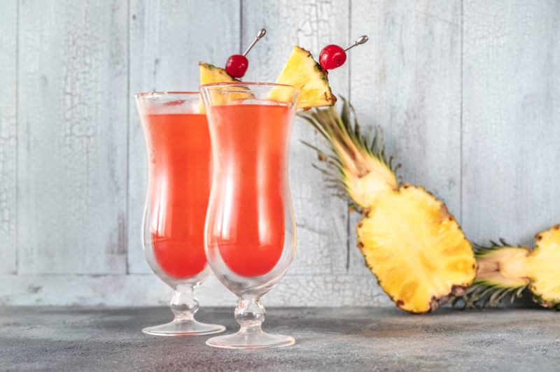 Two tall glasses of a Singapore sling cocktail in front of a wooden wall and a pineapple that's been sliced in half