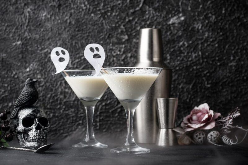 Two martini glasses containing banshee cocktails, each garnished with a paper figure of a ghost. There is a cocktail shaker and jigger next to them.