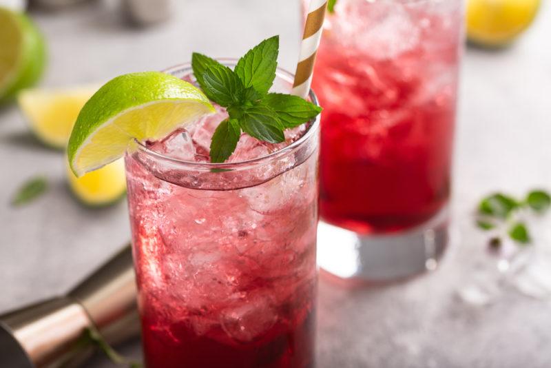 Two glasses of berry cocktail with lime wedges next to a cocktail jigger