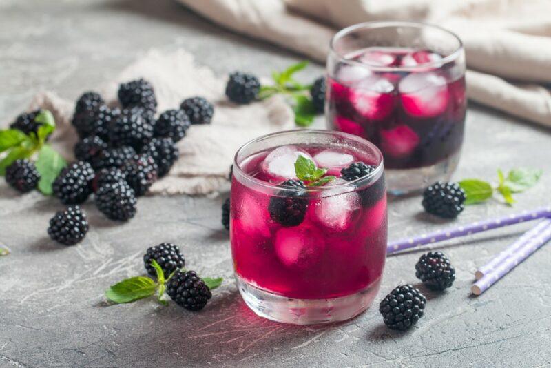 Two glasses of a blackberry Fireball whisky fizz cocktail with ice, with many blackberries on the table