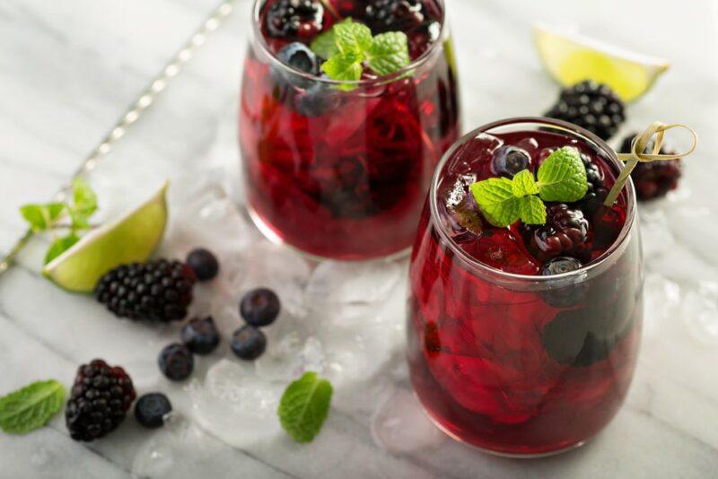 Two glasses containing a blackberry cocktail called goldsmith's daugher, which has been garnished with blackberries and blueberries