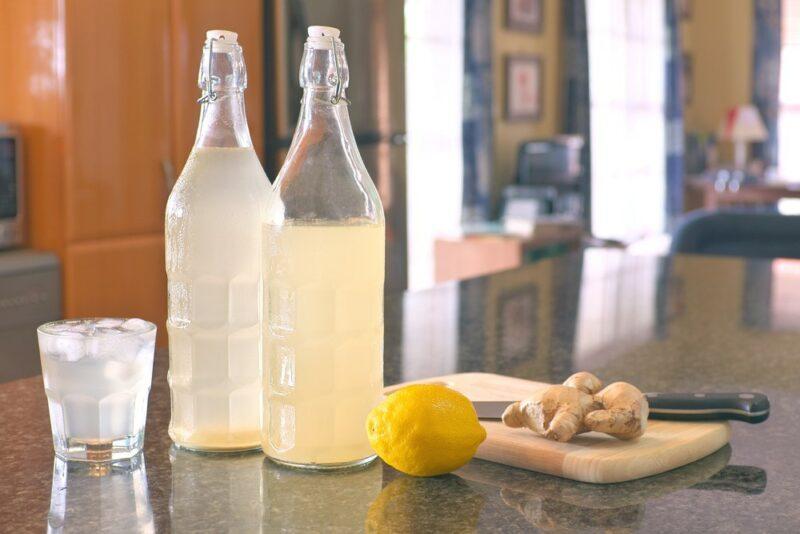 Two bottles of ginger ale and one glass of the ginger ale, next to a lemon and some ginger root, on a kitchen counter