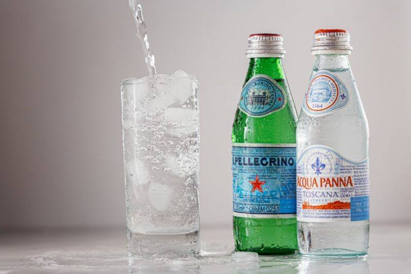 Two bottles of sparkling mineral water on a table next to a glass of sparking mineral water with ice and a straw