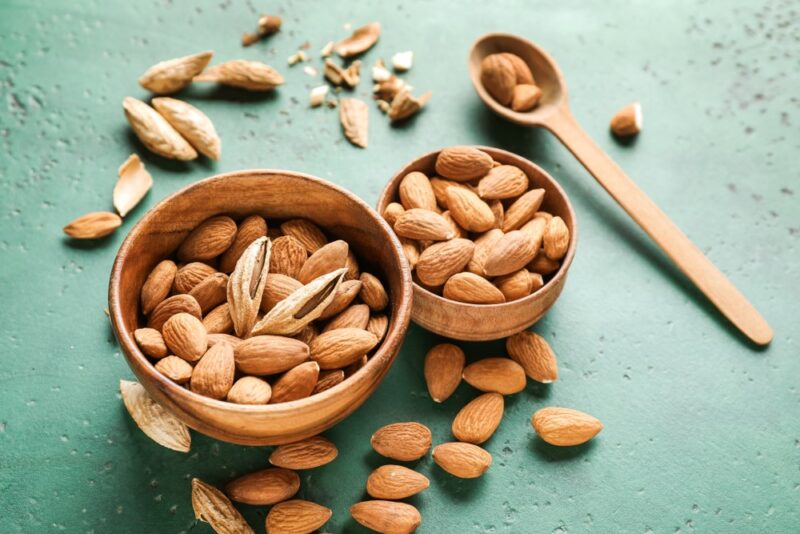 A light blue table with two bowls and a spoon of almonds, with more almonds scattered on the table