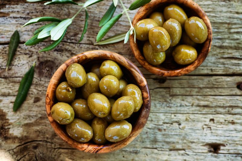 Two wooden bowls of green olives next to some olive branches on a wooden table