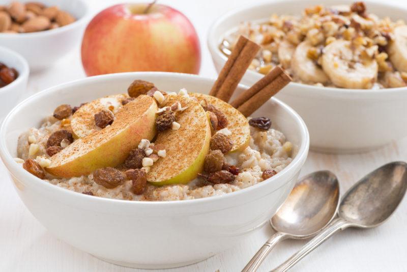 Two bowls of oatmeal with apples, raisins, and cinnamon, next to an apple, two spoons, and some small bowls of ingredients