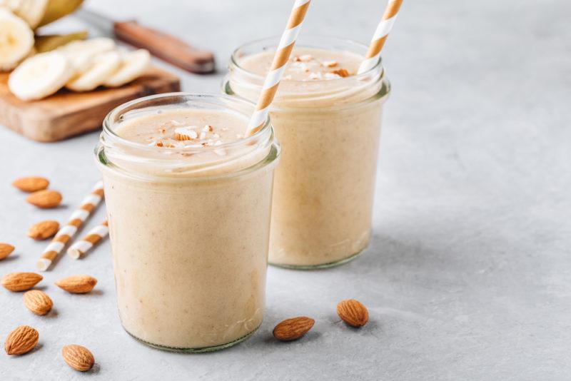 Two breakfast smoothies that include bananas and almonds in glasses with straws, with almonds and bananas on the table