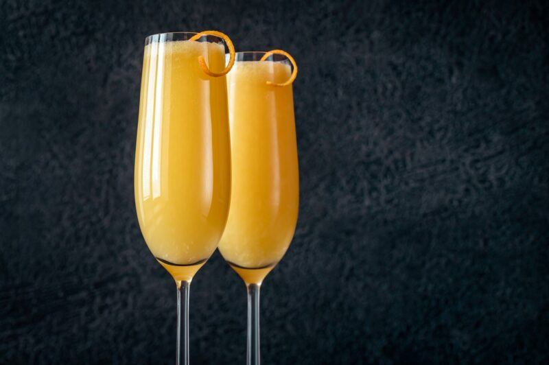 Two champagne glasses with a bucks fizz cocktail and a twist of citrus, in front of a black background