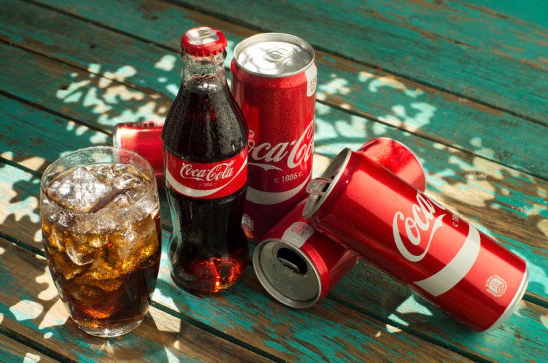 Cans of Coca Cola on a table, next to a bottle and a glass of the soda