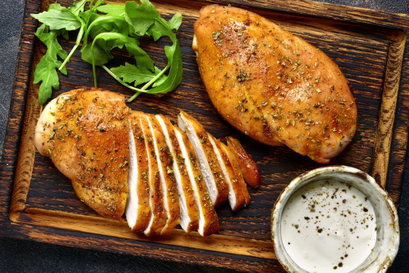 A wooden board with two chicken breasts, some herbs, and a small container of dip. ONe of the chicken breasts has been sliced