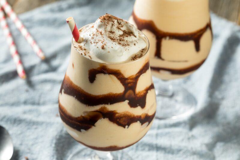 Two glasses of a chocolate mudslide dessert with layers of chocolate sauce and cream on top, with two straws on the table