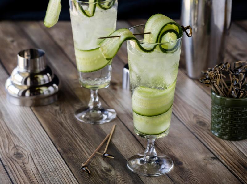 Two cocktails garnished with ribbons of cucumber on a wooden table in front of a bell