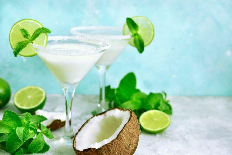 Two martini glasses containing coconut martinis, each garnished with a lime slice and some mint, next to mint, more limes, and half a coconut