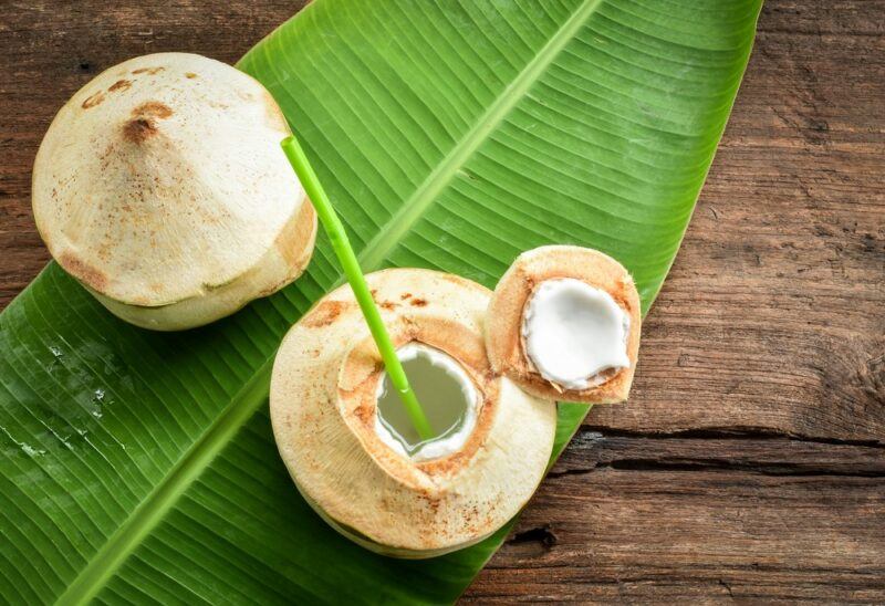 A wooden deck with a coconut leaf, one whole coconut, and one that has a straw sticking out of it