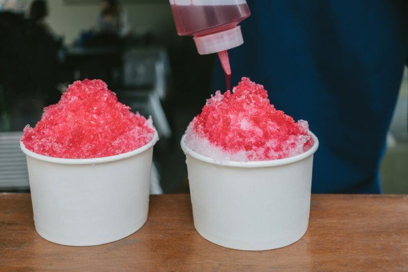 Two red tiger's blood snow cones in paper containers, where someone is pouring syrup over one of them