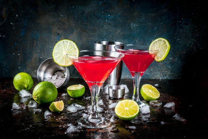 Two glasses of a cosmopolitan cocktail on a black table with lime wedges and ice as well