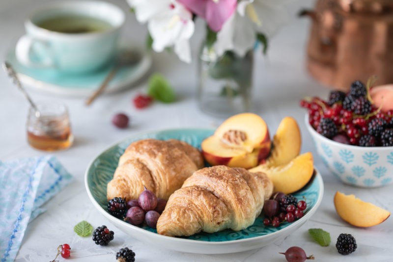 A light blue bowl with two croissants, peaches, and other fruit, with a mug of tea, a bowl of fruit, and various other things in the background