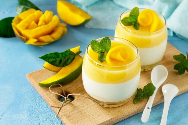 Two glasses of a yogurt and mango dessert, with mangos in the background