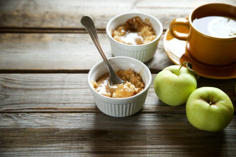 A wooden table with two ramekins containing a salted caramel apple crisp, a container of caramel and two green apples