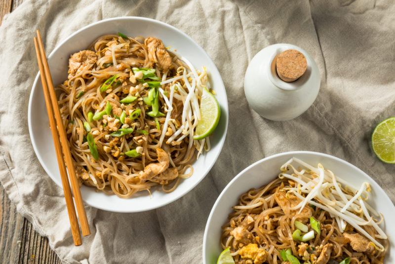 Two white dishes with pad thai, next to a white bottle, with two chopsticks on one of the dishes