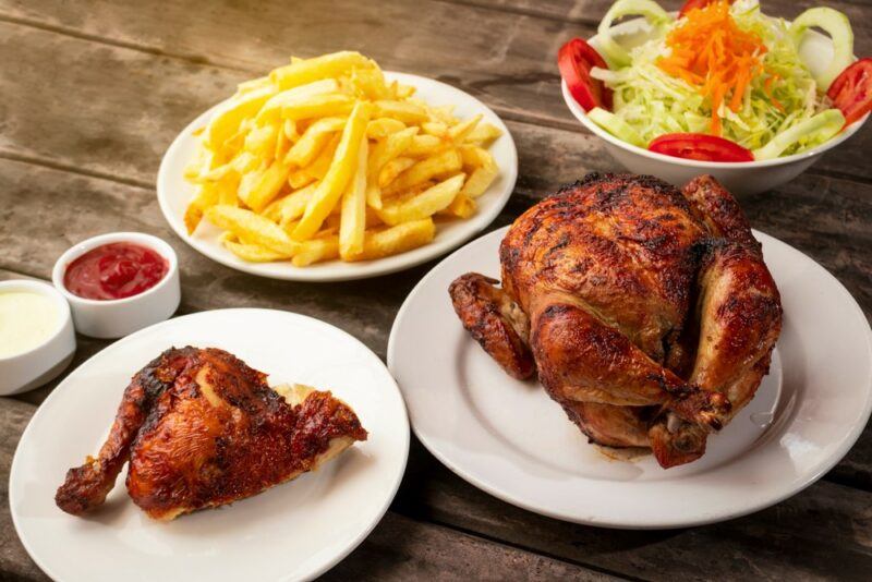 Two dishes of polla a la brasa on white plates, next to a plate of fries and another salad