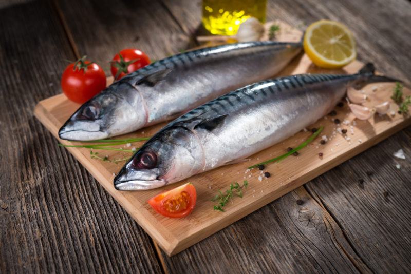 A cutting board with two fresh mackerel, tomatoes, and a lemon