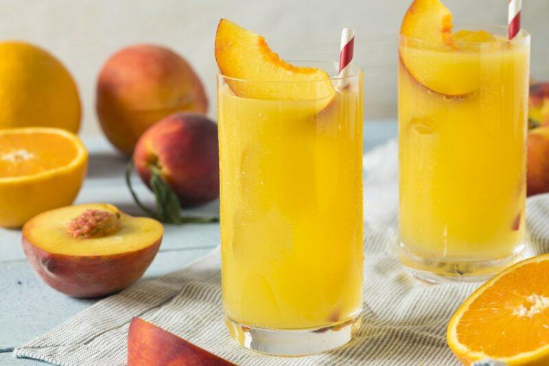 A wooden table with glasses containing fuzzy navel cocktails, each garnished with a piece of peach, with more fresh peaches and some oranges in the background