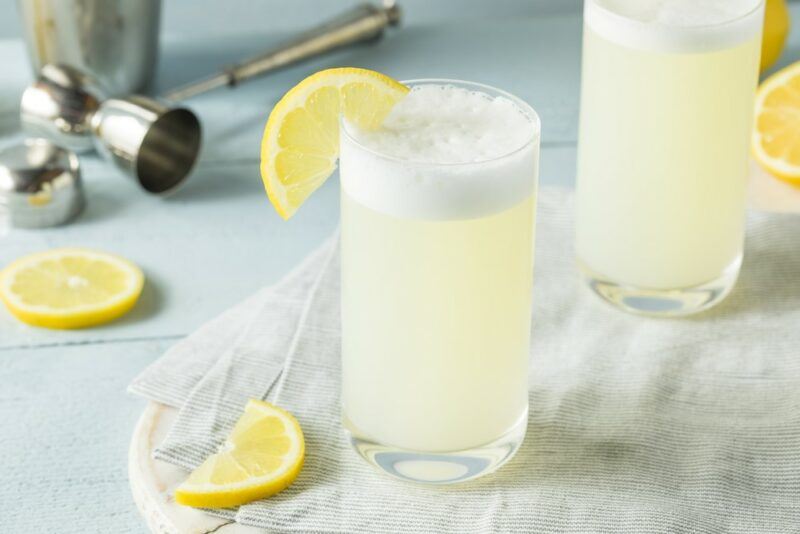 A gray board with two tall glasses of a gin fizz cocktail that are garnished with lemons. There are some lemon pieces on the table too