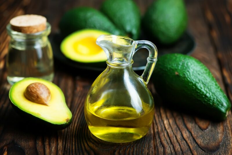 A wooden table with a bottle and jar of avocado oil, next to fresh avocados