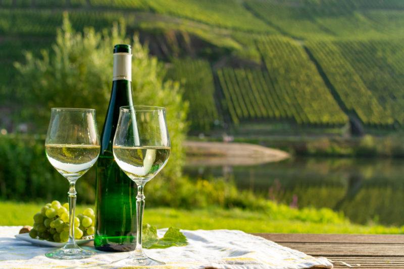 Two glasses and a bottle of reisling on a table next to some grapes, outside overlooking a vineyard