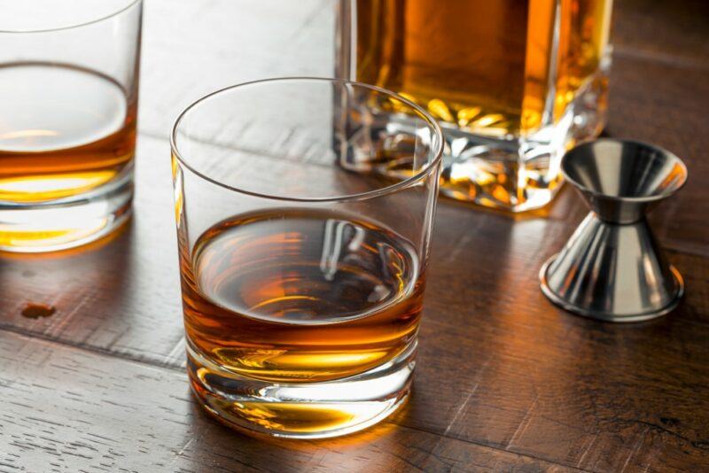 Two glasses of bourbon on a wooden table, next to a cocktail jigger and a decanter