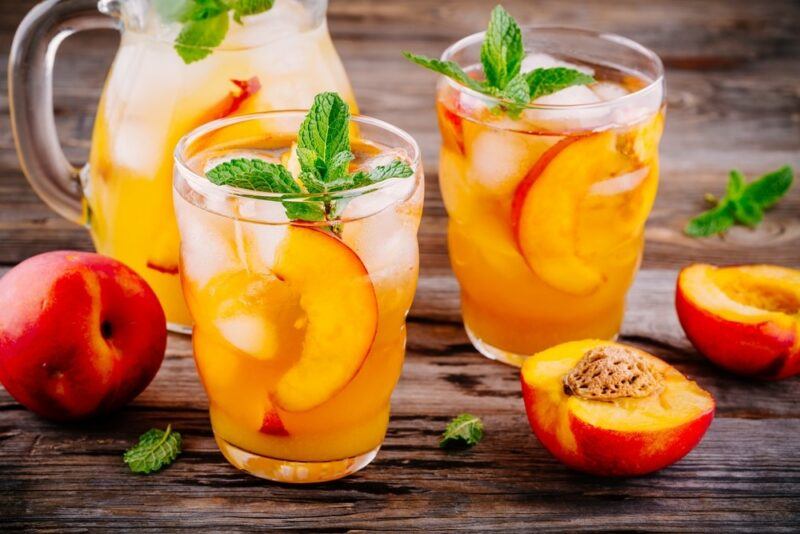 A wooden table with a jug and two glasses containing a peach cobbler moonshine cocktail, with sliced peaches and mint leaves