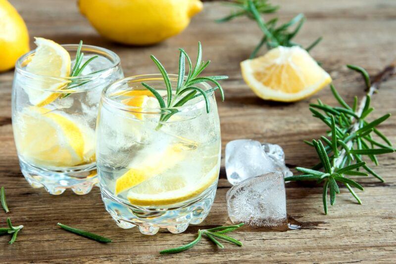 A wooden table with two glasses of gin and tonic, each garnished with rosemary, with ice, rosemary, and lemon on a table.