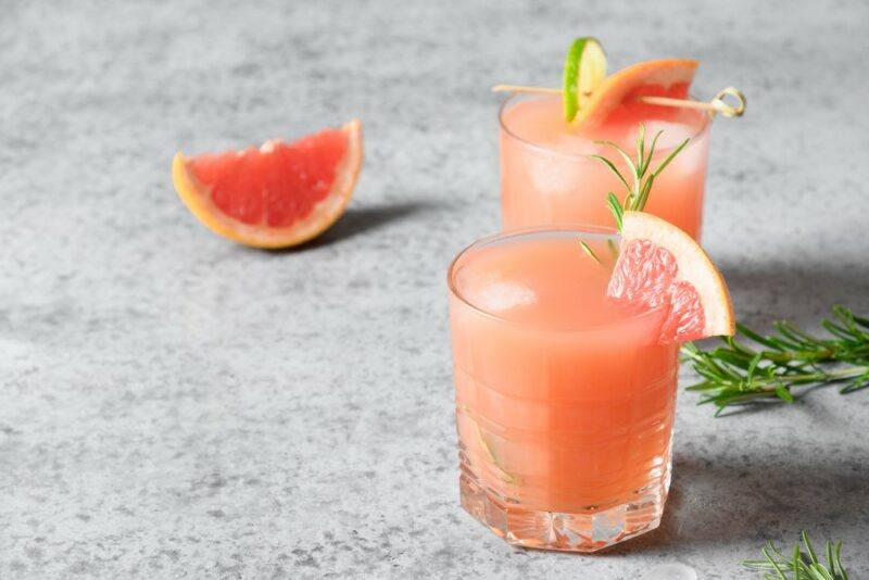 Two glasses containing a greyhound cocktail on a light gray table with a single wedge of grapefruit nearby