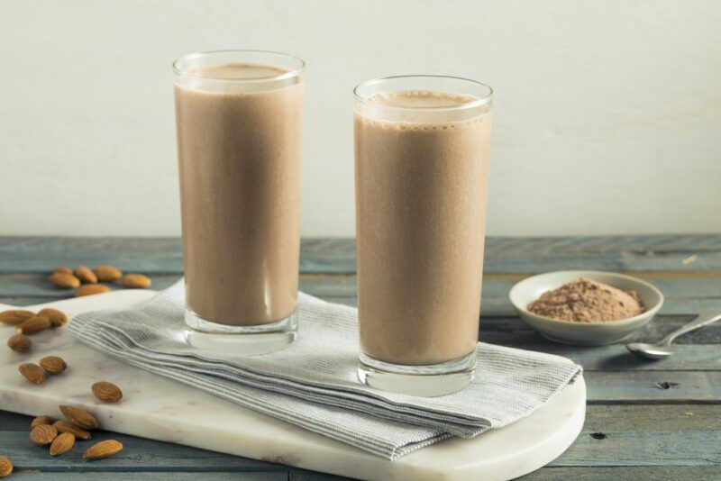 A wooden board with two glasses of chocolate protein shake, plus almonds, and a small dish of chocolate protein powder