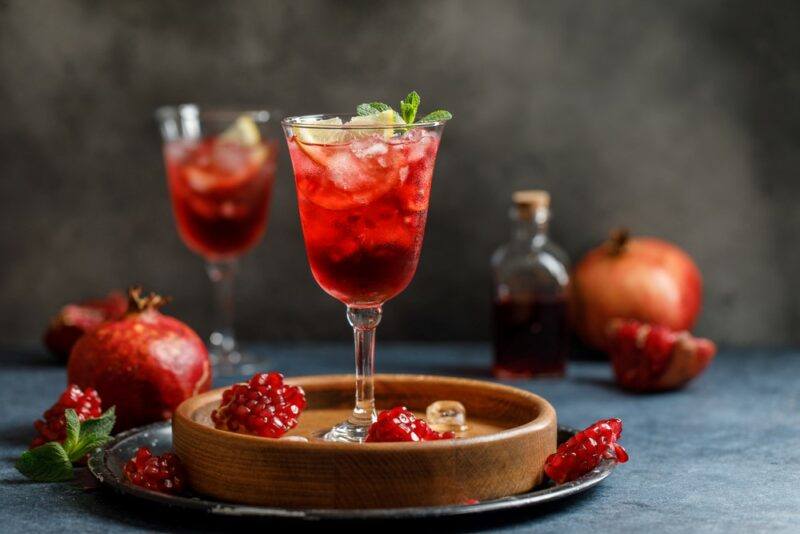 Two glasses containing a bright red cocktail called sakura smash, with pomegranates and pomegranate juice on the table