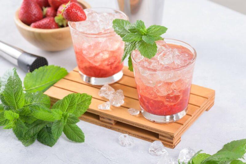 Two glasses containing a strawberry lemonade mojito on a wooden board, next to a bowl of strawberries and some mint