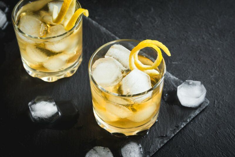 A black slate with two glasses of a whiskey mac cocktail with plenty of ice and a twist of lemon