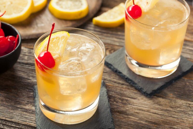 Two glasses of a whiskey sour cocktail with glace cherries, next to a bowl of the cherries and lemon slices