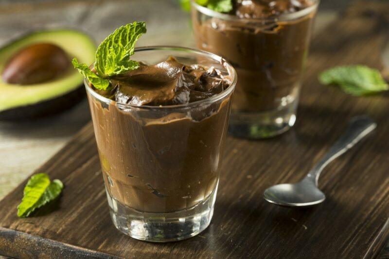 A wooden board with two glasses of avocado chocolate mousse, next to some mint leaves, a spoon, and an avocado