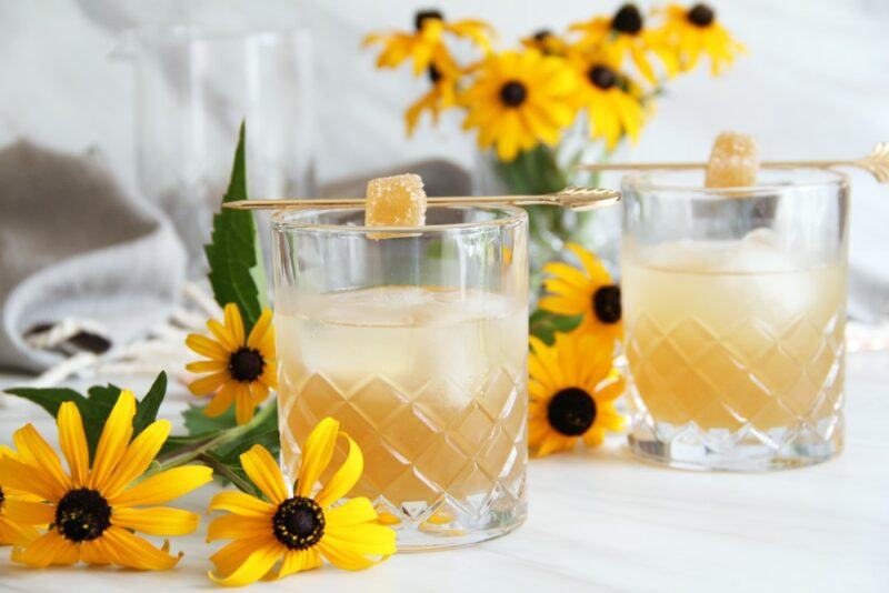 Two glasses of a bee's knees cocktail with ice and a sugar cube as a garnish, surrounded by yellow flowers